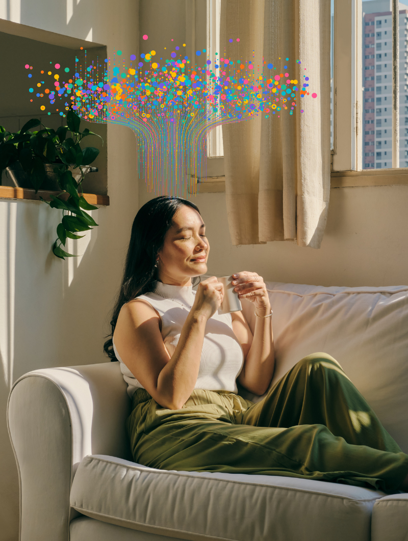 A woman sits comfortably on a couch, bathed in sunlight, with colorful dots and lines symbolizing thoughts of abundance emanating from her head towards the ceiling.